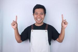 Smiling face expression young asian man wearing apron pointing up, empty space, isolated white background photo