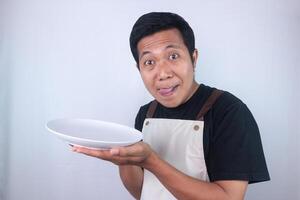 Funny expression asian young man showing delicious dish on white plate while stuck out his tongue. photo