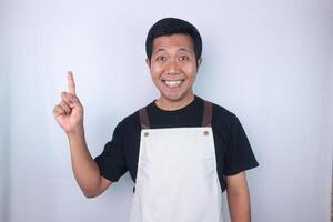 Smiling face expression young asian man wearing apron pointing up, empty space. photo