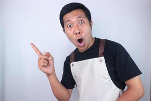 Funny shocked face expression young asian man wearing apron pointing right side, empty space. photo