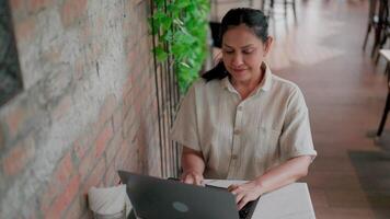 femme travail sur portable dans café. une âge moyen femme est assis à une table dans une café, concentré sur sa portable. elle est habillé mine de rien et apparaît à être travail ou en étudiant. video