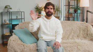 Happy young man vlogger smiling friendly at camera and waving hands while sitting on couch at home video