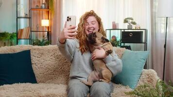 Young woman waving at call on smartphone with pug dog on home sofa, joyful friendly greeting video
