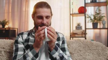 Young man drinking hot drink at home sofa, aroma, eyes closed, content sip, peaceful relaxation video