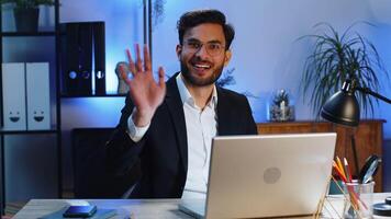 Businessman working on laptop smiling friendly at camera and waving hands gesturing hello at office video