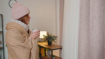 Young sick woman wear hat wrapped in plaid stay alone shivering from cold on sofa drinking hot tea video