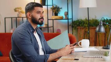 Indian freelancer man using smartphone texting message while staying in workplace at home office video