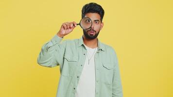 Indian young man holds magnifying glass near face looking at camera with big zoomed eye, analyzing video