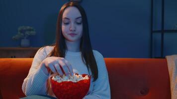 Woman sitting on sofa alone, eating popcorn and watching interesting tv serial, horror film at home video