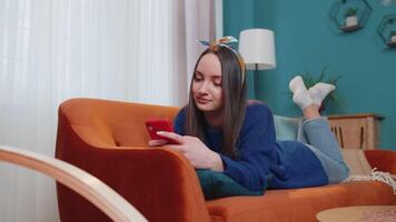 Joyful young adult woman lying on sofa using smartphone watching , online shopping, tapping video
