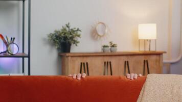 Man playing hide and seek peekaboo game hiding behind sofa looking at camera, positive smiling video