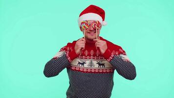 Funny man in red New Year sweater holding candy striped lollipops hiding behind them, fooling around video