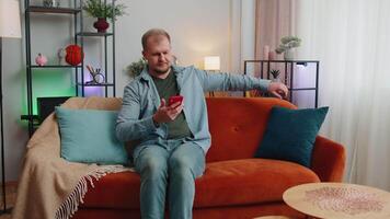 Cheerful young man sitting on sofa, using mobile phone share messages on social media application video