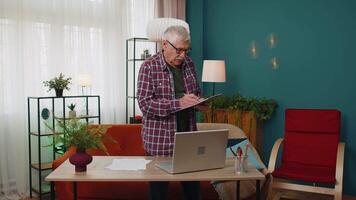 Senior grandfather man working on laptop computer making notes on sheet of paper calculating bills video