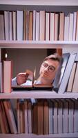 vertical Male researcher with eyeglasses immersed in reading an old book from a bookshelf in a quiet library at night, turning pages and diligently searching for valuable information video