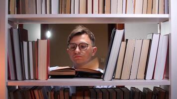 Young man wearing glasses is engrossed in reading a book, surrounded by towering bookshelves filled with a vast collection of literature, creating a cozy and intellectual atmosphere video