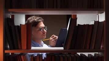 Young man wearing glasses writing notes on clipboard surrounded by books on shelves in a library, studying late at night with focused expression video