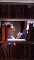 vertical Young man wearing glasses writing notes on clipboard surrounded by books on shelves in a library, studying late at night with focused expression video