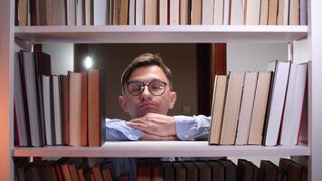 Young male librarian resting his chin on his hands, peering through a gap in a bookshelf filled with books, conveying a sense of peace and knowledge within a library setting video