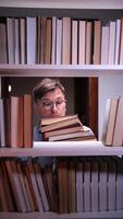 vertical Young student with eyeglasses peeking out from behind a bookshelf while clutching a stack of books in a university or public library, deeply engaged in the pursuit of knowledge video