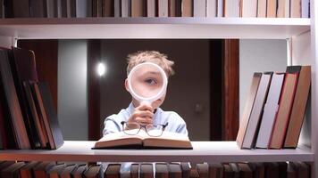 Young boy, possibly an elementary school student, holds a magnifying glass close to his eye while examining an open book in a library, surrounded by shelves filled with books video