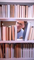 vertical Pensive student wearing glasses is searching for a book on a white bookshelf, resting his chin on his hand, surrounded by colorful books in a library or home setting video