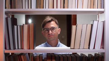 Young male librarian wearing eyeglasses, gazing thoughtfully between bookshelves in a library, surrounded by an impressive collection of books, embodying a passion for knowledge and learning video