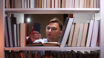 Man with glasses reading a book while using a smartphone in a library at night, surrounded by towering bookshelves filled with literature and knowledge video