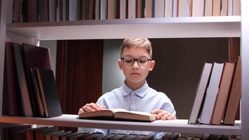 Concentrated pupil wearing eyeglasses reading a textbook sitting between bookshelves in a library, enjoying literature and studying for school video