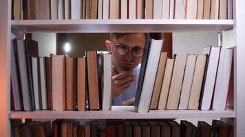 Pensive student wearing glasses is searching for a book on a white bookshelf, resting his chin on his hand, surrounded by colorful books in a library or home setting video