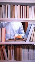 vertical Man in a blue shirt standing in front of a bookshelf, reading a book while holding a white mug, surrounded by an extensive collection of books, enjoying a moment of relaxation and focus video