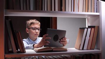 Curious young boy wearing eyeglasses using tablet computer for online education and reading book in home library at night, surrounded by bookshelves video