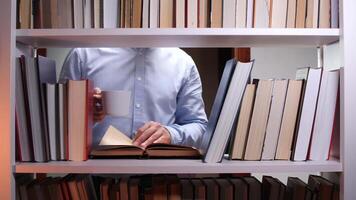 Man in a blue shirt standing in front of a bookshelf, reading a book while holding a white mug, surrounded by an extensive collection of books, enjoying a moment of relaxation and focus video