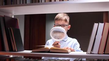 Concentrated young boy wearing eyeglasses using magnifying glass while reading interesting book standing between bookshelves in library, enjoying literature and gaining knowledge video