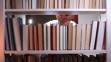 Thoughtful librarian with hand on chin is searching for a book among a vibrant array of colorful volumes on a white bookshelf in a serene library setting video