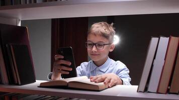 leende elev bär glasögon använder sig av smartphone och läsning lärobok medan Sammanträde mellan bokhyllor i bibliotek, njuter modern teknik för utbildning video