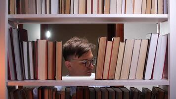 Young male librarian with eyeglasses searching for books on a shelf in a dimly lit library at night, carefully moving volumes with hands and intently looking between them video
