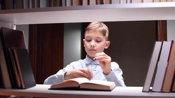 Concentrated pupil cleaning his eyeglasses with a microfiber cloth while sitting at a desk between bookshelves and reading a textbook, preparing for exams in a school library video