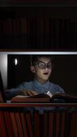 vertical Curious child in glasses is engrossed in reading a large book, illuminated by a flashlight, on a bookshelf filled with other books in a dimly lit room at night video