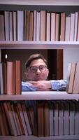vertical Young male librarian resting his chin on his hands, peering through a gap in a bookshelf filled with books, conveying a sense of peace and knowledge within a library setting video