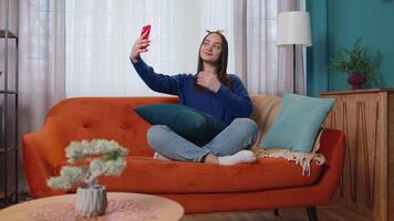 Woman on sofa at home with smartphone taking selfie on mobile phone cam, virtual call online video
