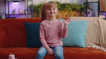 Child girl sitting on sofa at home looking at camera smiling waving hands gesturing hello or goodbye video