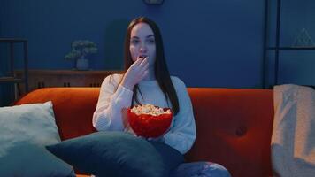 Woman sitting on sofa alone, eating popcorn and watching interesting tv serial, horror film at home video