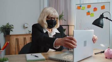 Senior employee wears face mask works alone from home office sits at desk using laptop computer video