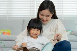 Happy Asian mother relax and read book with baby time together at home. parent sit on sofa with daughter and reading a story. learn development, childcare, laughing, education, practice. photo