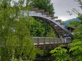 A modern footbridge spans a tranquil river, set against a backdrop of verdant trees and mountains. photo