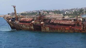 Aerial view of the Edro III shipwreck in Cyprus, partially submerged in blue waters near a rocky shoreline with houses, greenery, and coastal activity. video