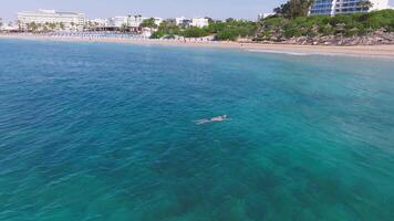 Aerial view of a lone swimmer in turquoise waters near Ayia Napa, Cyprus. White resorts line the coast, with a lighthouse visible on a jetty. video