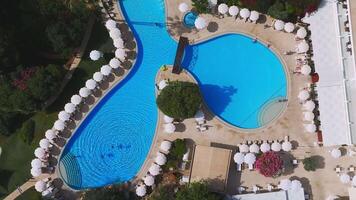 Aerial view of a resort in Ayia Napa, Cyprus, featuring a unique pool with a wooden bridge, white umbrellas, lounge chairs, and lush greenery. video