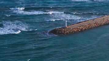 Aerial view of a white lighthouse on a stone jetty in Ayia Napa, Cyprus. Waves crash gently against the jetty, showcasing dynamic coastal motion. video